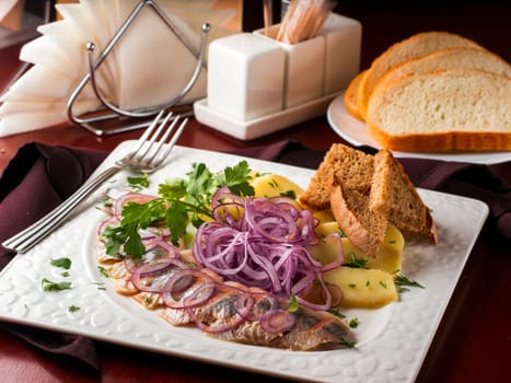 Appetizer of salted herring, red onion, potatoes and rye bread in white plate on dark brown wooden table in restaurant