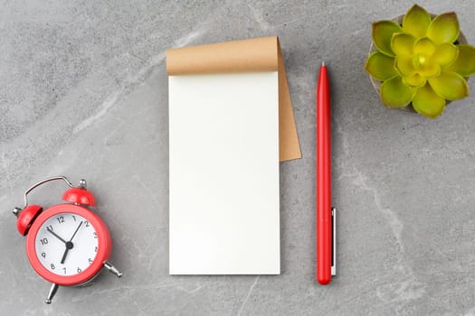 Open notepad with red pen, alarm clock and flower in pot on gray marble background. Flat lay