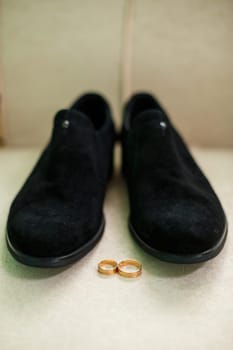 Gold wedding rings of the newlyweds lie next to men's shoes for the groom