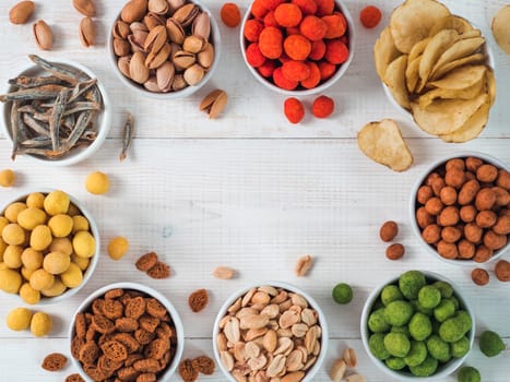 Assortment of different snack for beer, wine, party. Peanuts in coconut glaze, green vasabi, red spicy chilli, yellow cheese glaze, chips, pistachio, crackers, fish on white wooden table. Copy space