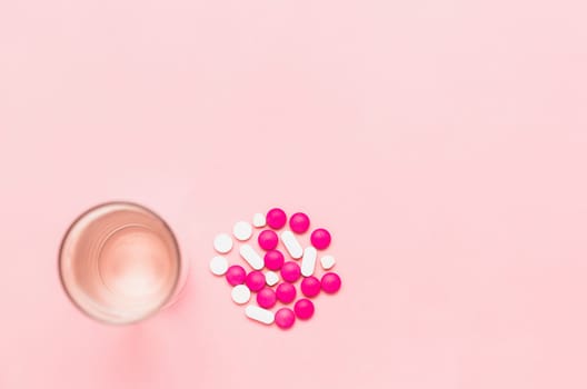 Top view of a glass of drinking water with piles of medicine pills on pink background for healthcare concept.