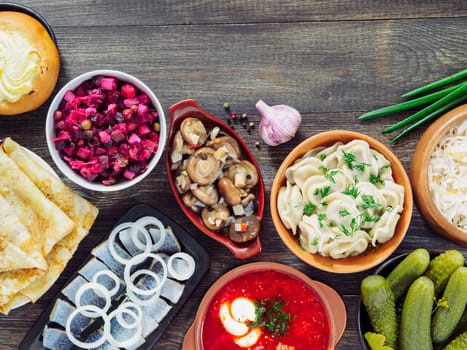 Russian food on wooden background. Assortment dishes of Russian cuisine - borscht, pelmeni, herring, marinated mushrooms, salted cucumbers, vinaigrette, sauerkraut and pancakes. Top view. Copy space.