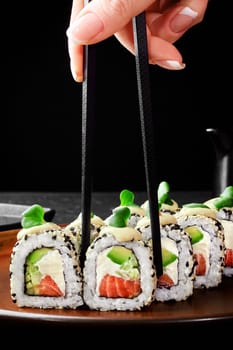 Appetizing Philadelphia rolls with salmon, cream cheese, avocado and cucumbers served on plate. Female hand taking one roll with chopsticks, cropped shot