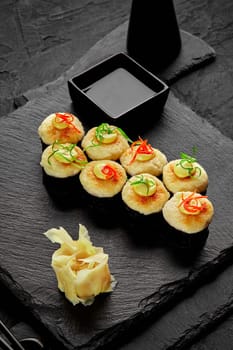 Baked norimaki with golden cheese caps topped with mayonnaise drops, colorful thin shaving of scallions and chili pepper traditionally served with pickled ginger and soy sauce on black slate board