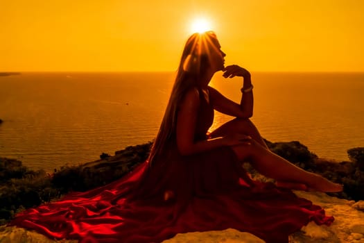 Woman sunset sea red dress, side view a happy beautiful sensual woman in a red long dress posing on a rock high above the sea on sunset