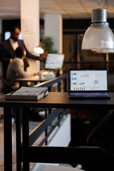Laptop computer with financial graphs on screen, standing on table in startup office, coworkers discussing company strategy in background. Employees working late at night in startup office