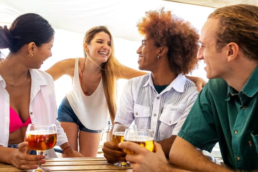 Young blond woman talking to friends in beach bar while having drinks together. Multiracial friends having fun. Summertime and vacation concept.