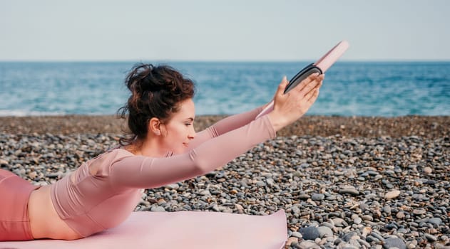 Middle aged well looking woman with black hair doing Pilates with the ring on the yoga mat near the sea on the pebble beach. Female fitness yoga concept. Healthy lifestyle, harmony and meditation.