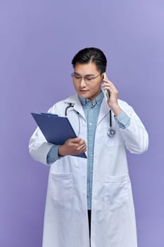 Attractive young cheerful male doctor wearing uniform taking notes while talking on mobile phone