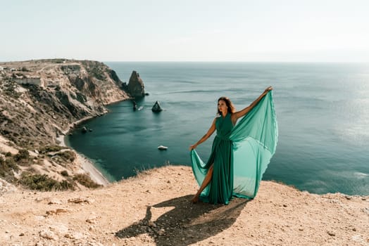 Woman green dress sea. Female dancer posing on a rocky outcrop high above the sea. Girl on the nature on blue sky background. Fashion photo