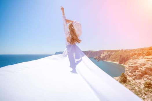 woman sea white dress. Blonde with long hair on a sunny seashore in a white flowing dress, rear view, silk fabric waving in the wind. Against the backdrop of the blue sky and mountains on the seashore