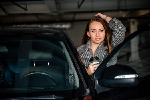 Happy woman car. she stands next to the car in the underground parking. Dressed in a gray coat, holding a glass of coffee in her hands, a black car