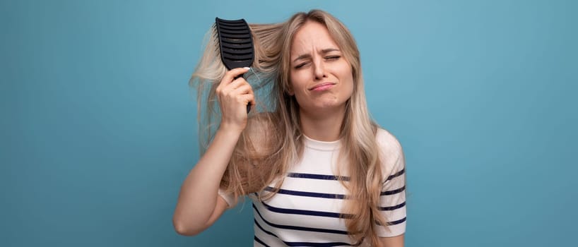 horizontal photo of a sad upset blond young woman with a hairbrush in her hands on a blue background.