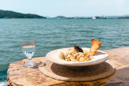 Seafood pasta spaghetti and a glass of water standing on a wooden table in outside cafe restaurant with scenery sea in the background. Copy blank space. Fish diving into noodles concept.