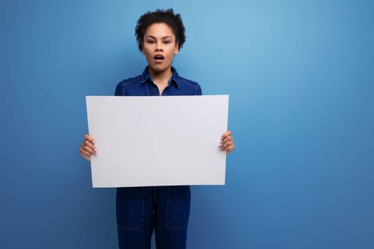 young pretty hispanic business woman with fluffy hair dressed in blue denim suit holding billboard with blank space.