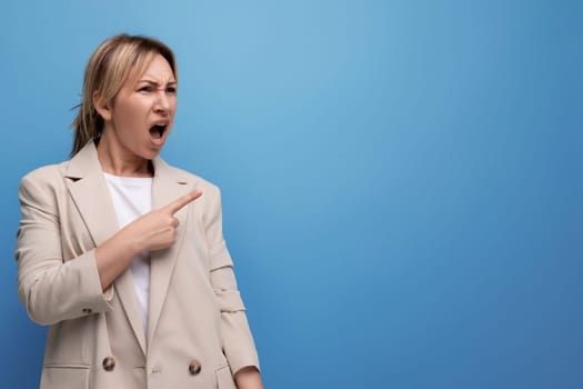 surprised blond young woman in jacket pointing finger to side on studio background.