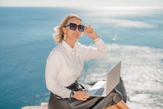 Business woman on nature in white shirt and black skirt. She works with an iPad in the open air with a beautiful view of the sea. The concept of remote work