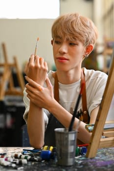 Portrait of young Asian gay man wearing apron holding paintbrush sitting in art workshop. Hobby and leisure concept.