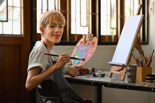Young hipster queer artist holding palette and brush sitting in front of easel in bright studio. Leisure activity and art concept.