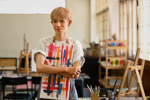 Portrait of pretty young gay man in apron holding art canvas and smiling to camera. Leisure activity, creative hobby and art concept.