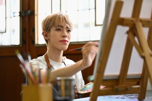 Pleased young gay man painting with watercolor on canvas in art workshop. Art, creative hobby and leisure activity concept.