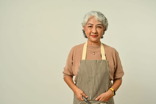 Positive grey haired woman 50s wearing apron standing isolated on gray background. People, household, small business owner concept.