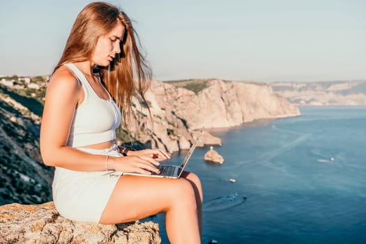 Successful business woman in yellow hat working on laptop by the sea. Pretty lady typing on computer at summer day outdoors. Freelance, travel and holidays concept.