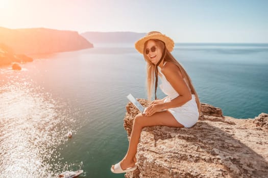 Successful business woman in yellow hat working on laptop by the sea. Pretty lady typing on computer at summer day outdoors. Freelance, travel and holidays concept.