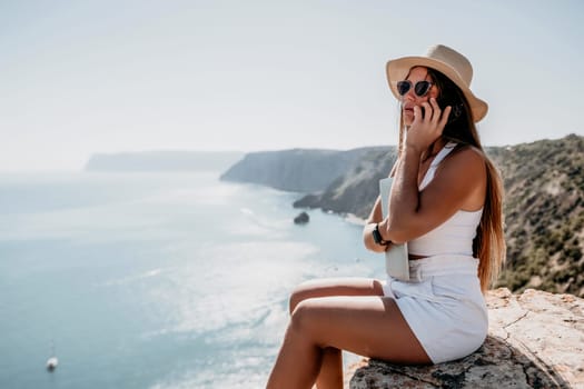 Successful business woman in yellow hat working on laptop by the sea. Pretty lady typing on computer at summer day outdoors. Freelance, travel and holidays concept.