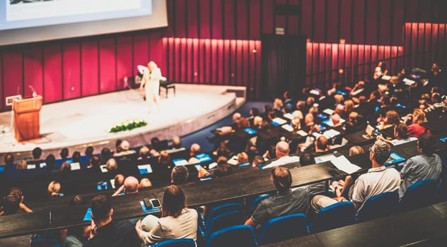 Business and entrepreneurship symposium. Female speaker giving a talk at business meeting. Audience in conference hall. Rear view of unrecognized participant in audience.