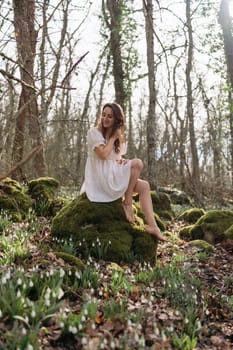 Portrait of a woman in the forest. She is sitting in a white dress on a meadow with snowdrops in a spring forest.