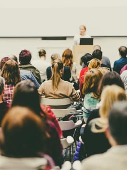 Business and entrepreneurship symposium. Female speaker giving a talk at business meeting. Audience in conference hall. Rear view of unrecognized participant in audience. Copy space on whitescreen.