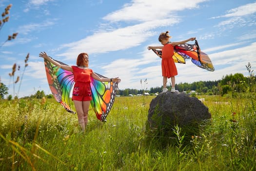 Happy female family with red haired mother and daughter with bright butterfly wings having fun on green and yellow meadow full of grass and flowers in sunny summer day. Concept family love