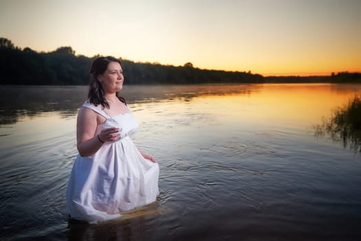 Slavic plump plump chubby girl in long white dress on the feast of Ivan Kupala with flowers and water in a river or lake on summer evening