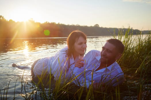 Beautiful adult couple having hugs and fun on nature in the water of a river or lake in the summer evening at sunset. A guy and a girl swim and relax outdoors in clothes in white shirts and jeans