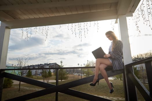 Beautiful blonde girl working by laptop in gazebo on an autumn, summer or spring day. Businesswoman, student, freelancer or manager works outdoors in natural landscape in village, city or town