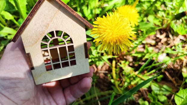 Small wooden toy house on palm of woman hand on natural background with yellow dandelion. concept of care, buying, selling, donating of eco friendly home. copy space. close-up. soft selective focus