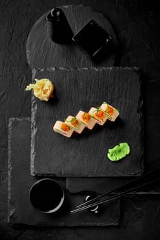 Set of sushi rolls wrapped in mamenori topped with tobiko and shavings of scallions traditionally served with wasabi, gari and soy sauce on slate board, top view on black background. Japanese food