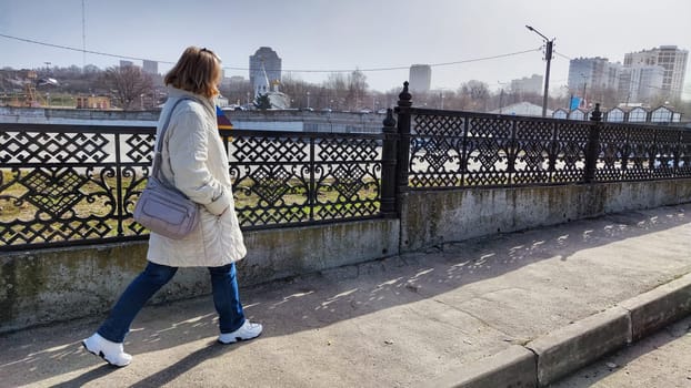 A focused, thoughtful woman in a warm light jacket, jeans and sneakers is walking briskly with wide steps along a street with a lace fence on a sunny day