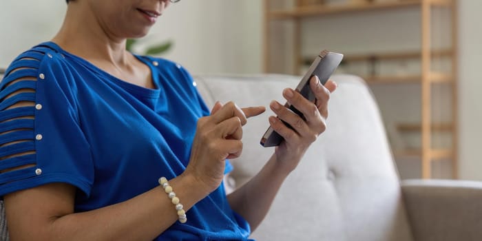 Smiling older asian woman sit rest on couch at home text or message online on modern cellphone gadget. Happy mature female relax browse internet on smartphone device. Elderly technology concept.