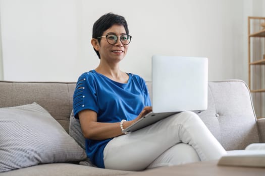 Happy successful mature business woman elderly working from home, holding laptop, looking at camera, smiling, resting in armchair, using modern technology, wireless connection for remote job.