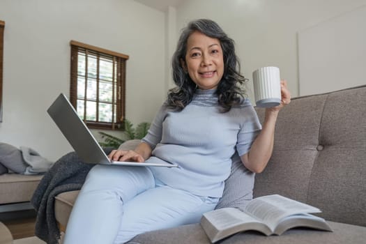 Pleasant chat. Beautiful elderly woman texting on her laptop and smiling happily while drink coffee sitting on the couch in the living room.