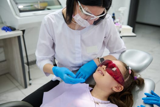 Caucasian experienced pleasant woman dentist doctor, dental hygienist talks to a little patient, adorable little child girl sitting in dentist's chair during appointment in pediatric dentistry clinic