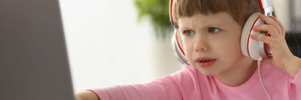 Little girl child in headphones in front of laptop. Children emotions of discontent and focusing attention concept