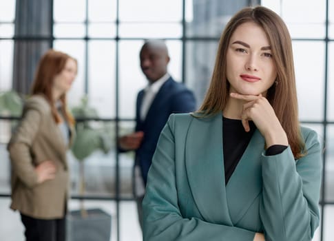 businesswoman standing with her arms crossed in an office while her colleagues have a meeting in the background.