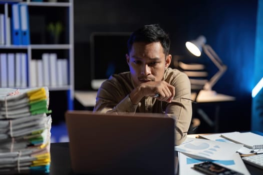 Overworked young Asian office employee working on laptop computer overtime in office at night.