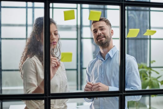 Creative professionals standing and discussing in office behind glass wall with sticky notes