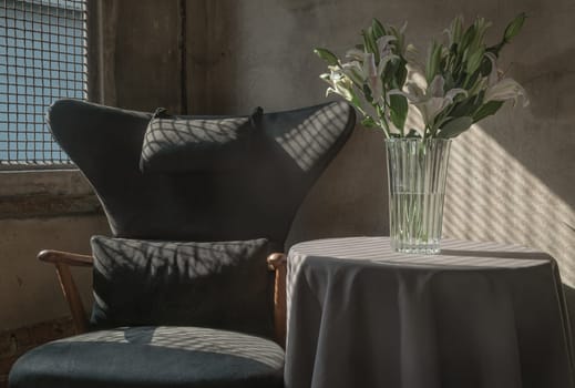 A Wooden Upholstered Chair and the Bouquet of Flowers in a Glass Vase on Table in the Room with Bare Cement Wall. They are affected by the Sunbeam Shining through Steel Mesh Window in the Afternoon. Space for text, Selective focus.