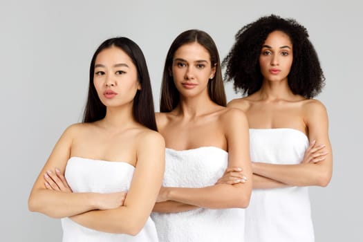 Diverse beauty. Portrait of three beautiful multiracial women posing with folded arms wrapped in white towels and smiling at camera while standing in line over grey background