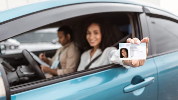 Passed Exam. Happy middle eastern woman sitting in car showing driver license from window, celebrating driving school finish, inside of vehicle with instructor man on passenger seat. Focus on card
